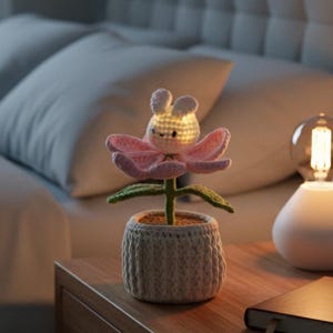 May include: A crocheted flower night light with a bunny head center, pink petals, and green leaves. The flower sits in a gray crocheted pot on a wooden nightstand. The light is on, illuminating the bunny's face.