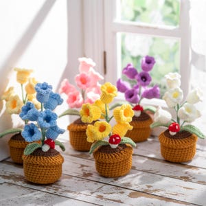 May include: A collection of handmade crochet flowers in small brown pots. The flowers are in various colors, including yellow, blue, pink, purple, and white. Each pot has a small mushroom decoration. The flowers are arranged on a wooden surface.