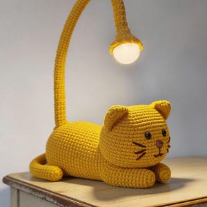 May include: A yellow crochet cat lamp with a curved neck and a round light. The cat has black eyes, a brown nose, and whiskers. The lamp is sitting on a light-colored wooden table.