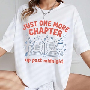 Può includere: Una t-shirt bianca a maniche corte con una stampa grafica che riporta il testo "JUST ONE MORE CHAPTER" e "up past midnight" in rosso. Il design include un'illustrazione a linee blu di un libro aperto, una tazza di caffè, fiori e stelle.