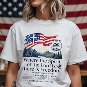 Könnte beinhalten: Ein weißes T-Shirt mit einer Grafik einer wehenden amerikanischen Flagge, einem Kreuz, einem Siegel zum 250-jährigen Jubiläum und Bergen. Der Text lautet: Where the Spirit of the Lord is, there is Freedom, 2 Corinthians 3:17, Faith, Freedom, Grace, Independence Day 2026.