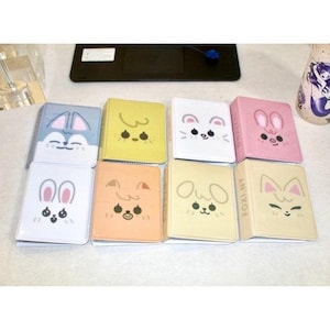 May include: A selection of small, colourful notebooks featuring cartoon animal faces. The notebooks are in various colours, including blue, yellow, white, pink, and beige. Each notebook has a different animal design, such as a bunny, dog, or cat.