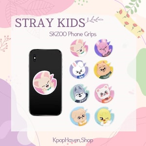 Peut inclure: Un smartphone noir avec un support de téléphone représentant un personnage de dessin animé rose et blanc. L'image présente également plusieurs autres supports de téléphone SKZOO avec différents designs de personnages. Le texte sur l'image indique "STRAY KIDS" et "SKZOO Phone Grips."