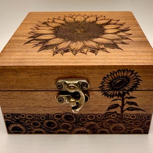 May include: A wooden box with a sunflower design. The top of the box features a large sunflower engraving, while the front has a smaller sunflower and a field of sunflowers engraved along the bottom. The box has a metal clasp.