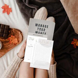 Könnte beinhalten: Ein graues Notizbuch mit dem Text "Manage Your Money" und "Monatsübersicht" auf dem Cover. Das Notizbuch ist auf einer Seite mit einem Monatskalender und einer Seite mit einer Liste zur Verfolgung von Ausgaben geöffnet.