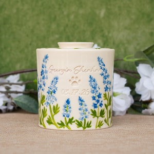 May include: A cylindrical cream-coloured ceramic urn featuring a hand-painted pattern of blue lupine flowers with green stems. The front is engraved with the name Georgie Sheehan, a paw print icon, and the date 03.17.26 in a simple script font.