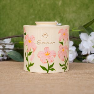 May include: A cylindrical cream-coloured ceramic container featuring a painted design of pink lilies with green stems and leaves. The word Summer is written in cursive script above the central flower, accompanied by a small sun icon above the text.