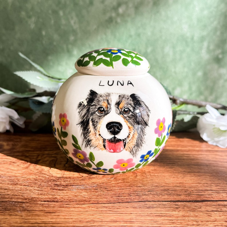 May include: A white ceramic pet urn with a hand-painted portrait of a dog. The urn is decorated with colorful flowers and the name "LUNA" is written on the front. The lid is also decorated with flowers.