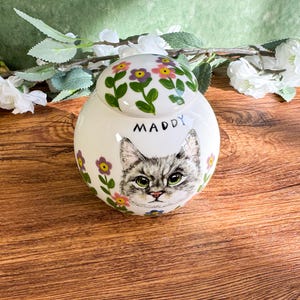 May include: A white ceramic jar with a lid, featuring a hand-painted gray cat portrait with green eyes and the name "Maddy". The jar is adorned with floral designs in pink, yellow, and purple, with green leaves. The jar is approximately 6 inches tall.