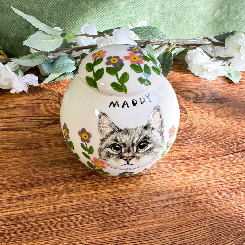 May include: A white ceramic jar with a lid, featuring a hand-painted gray cat portrait with green eyes and the name "Maddy". The jar is adorned with floral designs in pink, yellow, and purple, with green leaves. The jar is approximately 6 inches tall.