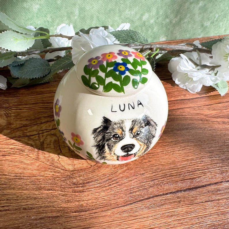 May include: A cream-colored ceramic urn with a painted portrait of a dog and the name "LUNA". The lid is decorated with colorful flowers. The urn is a memorial item for pets.