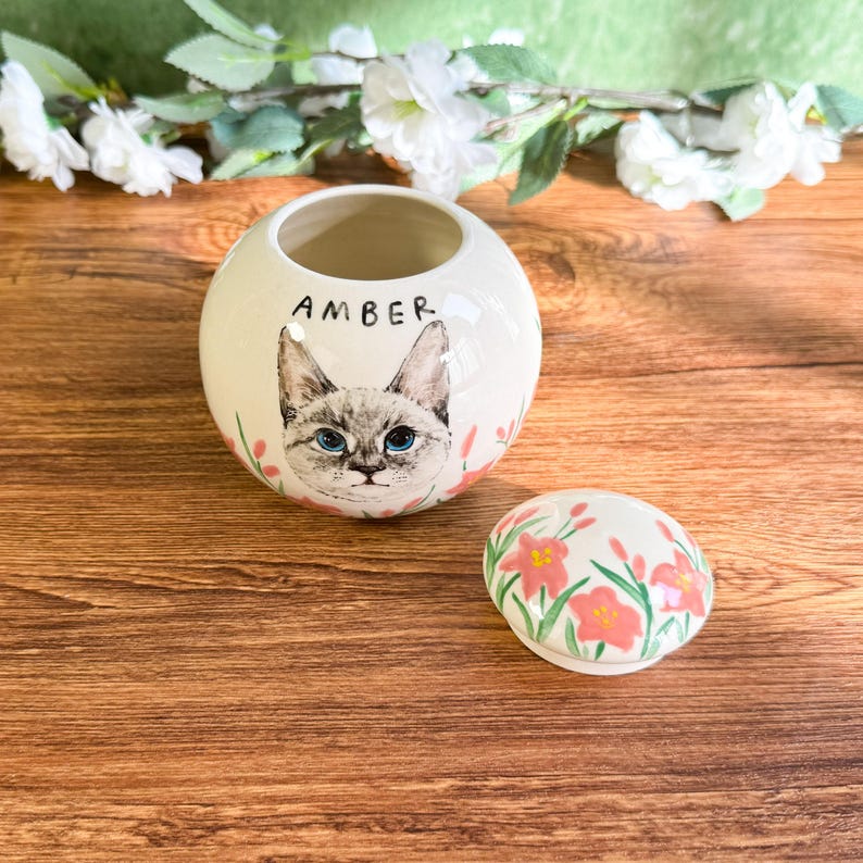 May include: A white ceramic jar with a cat face illustration and the name "AMBER" in black lettering. The jar has a matching lid with pink floral designs. The cat has blue eyes and is surrounded by pink flowers.