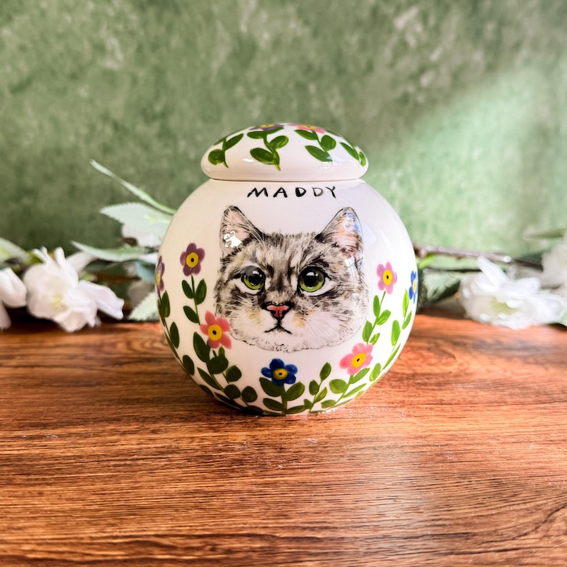 May include: A white ceramic pet urn with a hand-painted portrait of a gray tabby cat. The urn is decorated with colorful flowers and green leaves. The name "Maddy" is written above the cat's portrait. The lid is also decorated with flowers and leaves.