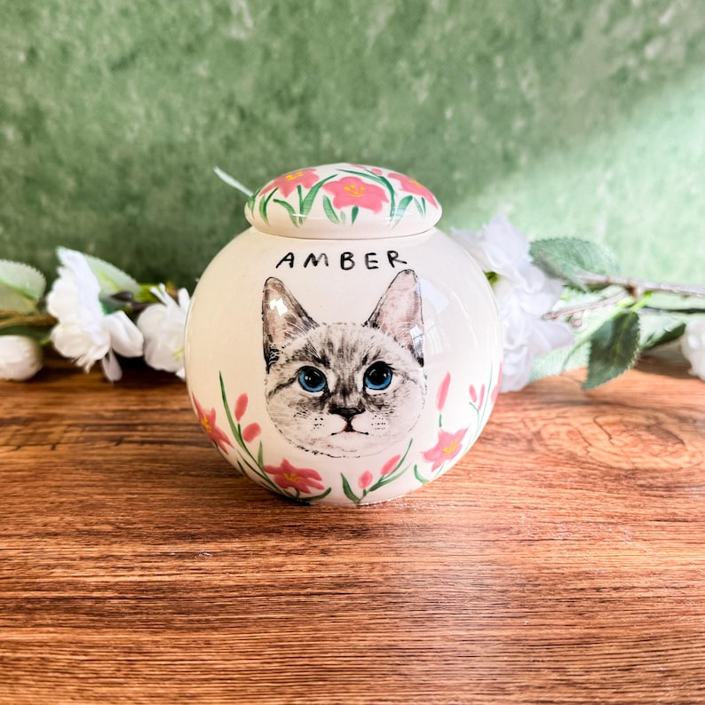 May include: A white ceramic pet urn with a hand-painted cat portrait and the name "AMBER" in black. The urn is decorated with pink flowers and has a matching lid. The cat has blue eyes.