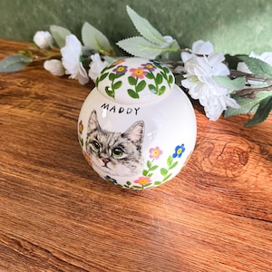 May include: A white ceramic pet urn with a hand-painted portrait of a gray tabby cat named Maddy. The urn is decorated with colorful flowers and has a matching lid with floral accents. The urn is a round shape.