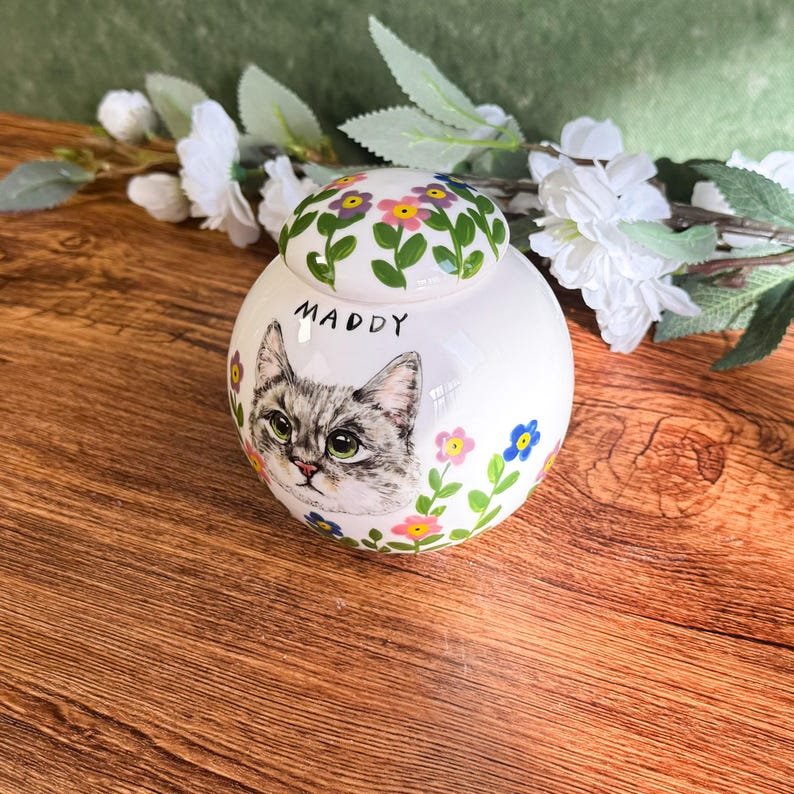 May include: A white ceramic pet urn with a hand-painted portrait of a gray tabby cat named Maddy. The urn is decorated with colorful flowers and has a matching lid with floral accents. The urn is a round shape.