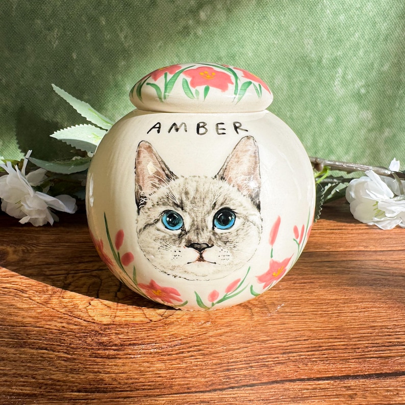 May include: A round, off-white ceramic urn with a hand-painted cat portrait and the name "AMBER". The urn features a lid adorned with pink flowers and green leaves. Pink flowers and green leaves are also painted around the cat portrait.