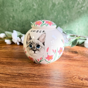 May include: A white ceramic pet urn with a hand-painted cat portrait and the name "AMBER". The urn is decorated with pink flowers and a matching lid. The cat has blue eyes and gray and white fur.