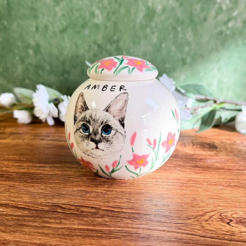 May include: A white ceramic pet urn with a hand-painted cat portrait and the name "AMBER". The urn is decorated with pink flowers and a matching lid. The cat has blue eyes and gray and white fur.