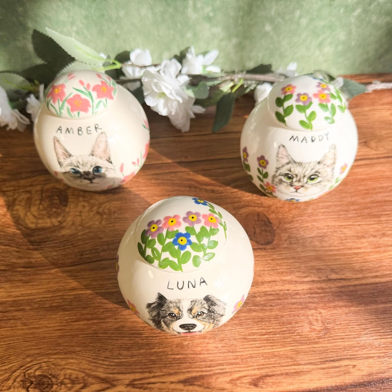 May include: Three ceramic pet urns with floral lids. Each urn features a painted portrait of a cat or dog, with the pet's name below. The urns are white with colorful floral accents.