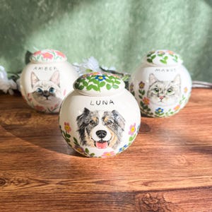 May include: Three ceramic pet urns with hand-painted portraits of a cat and a dog. The urns are white with floral accents and the names "Amber", "Luna", and "Margot". The lids are decorated with flowers and green leaves.
