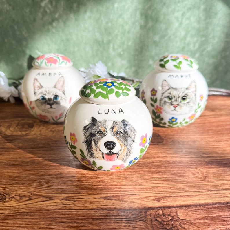 May include: Three ceramic pet urns with hand-painted portraits of a cat and a dog. The urns are white with floral accents and the names "Amber", "Luna", and "Margot". The lids are decorated with flowers and green leaves.
