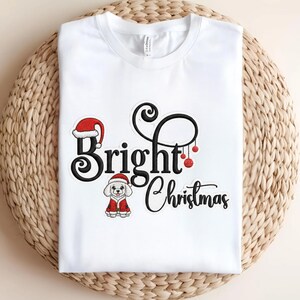 Könnte beinhalten: Weißes T-Shirt mit einem festlichen "Bright Christmas" Design. Das Design beinhaltet einen Weihnachtsmannhut, einen kleinen Hund in einem Weihnachtsmannanzug und rote Kugeln. Das Shirt ist auf einer gewebten Oberfläche gefaltet.