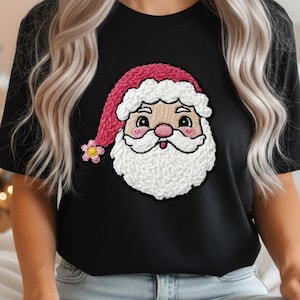 May include: Black t-shirt featuring an embroidered Santa Claus face. The Santa design includes a pink hat, white beard, and rosy cheeks. A small flower is on the hat. The shirt is made of a soft material.