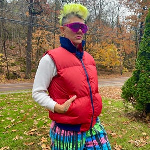 May include: A person wearing a red puffer vest with a navy blue collar and zipper, over a white long-sleeved shirt. They are also wearing colorful patterned pants and pink sunglasses. The background features a forest with autumn foliage.