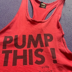 May include: A red tank top with the words "PUMP THIS!" printed in dark brown. The tank top has a racerback design and a small logo near the bottom. The shirt is made of a soft material.