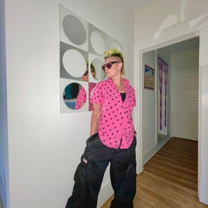May include: A person wearing a pink button-down shirt with black polka dots, black sunglasses, and black cargo pants. The person has a mohawk hairstyle and is standing in front of a white wall with a circular mirror.