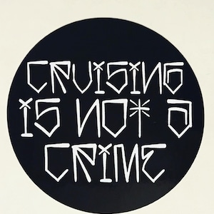 May include: A round black sticker with white text that reads "CRUISING IS NOT A CRIME". The text is stylized with geometric shapes. The sticker is likely for a car or other vehicle.