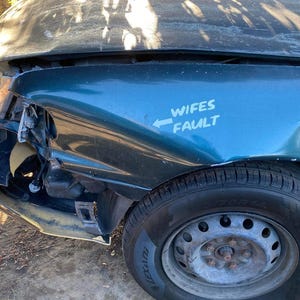 May include: A damaged teal car with a dented front fender and a flat tire. The front end is heavily damaged, with the headlight and bumper broken. The text "Wifes Fault" is written on the fender.