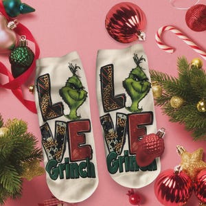 May include: A pair of white socks with the word "LOVE Grinch" printed on them. The letters "L" and "V" have a leopard print design. The Grinch's face is green. Christmas-themed socks.