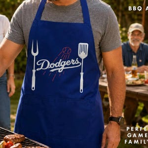 May include: A royal blue BBQ apron featuring a white graphic of a baseball, a two-pronged meat fork, and a spatula. The text Dodgers is printed in white script in the center. The apron includes a front pocket and neck strap.