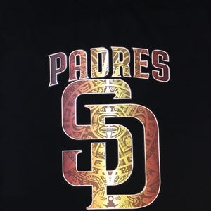 Puede incluir: Camiseta negra con un logotipo de los San Diego Padres en dorado y rojo. El logotipo presenta una "SD" con un diseño de inspiración azteca.