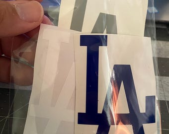 May include: A clear plastic bag containing several white vinyl stickers. The stickers feature the blue and white "LA" logo, with a gray "T" sticker visible. The stickers are likely for application on various surfaces.
