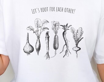 Può includere: T-shirt bianca con la scritta "Let's Root For Each Other!" e un'illustrazione in bianco e nero di varie verdure a radice. La maglietta è a girocollo.