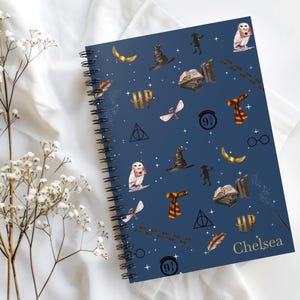 Puede incluir: Un cuaderno con espiral de color azul oscuro con un patrón repetido de elementos mágicos dorados, como alas y búhos. El nombre "Chelsea" está impreso en dorado en la portada.