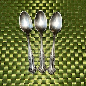 May include: Three silver-toned metal spoons with an oval bowl and a decorative, ornate handle featuring intricate embossed scrollwork patterns. The spoons have a polished finish with visible signs of ageing and patina along the metal surfaces.