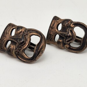 May include: Pair of antique-style cufflinks featuring a bronze finish. Each cufflink is shaped like a theatrical mask, with a detailed design of a smiling face. The cufflinks are likely made of metal and are set against a white background.