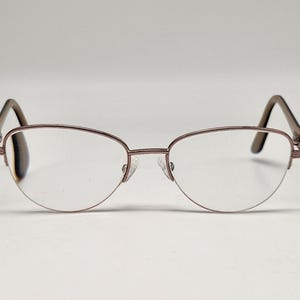 May include: Semi-rimless eyeglasses featuring a metallic rose gold frame with a textured finish on the bridge and temples. The lenses are clear and oval-shaped, supported by adjustable nose pads and dark brown temple tips for a secure fit.