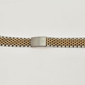 May include: A two-tone watch band featuring a multi-link design with alternating silver-colored and gold-colored metal segments. The bracelet includes a central rectangular metal clasp mechanism that connects the two sides of the watch strap.