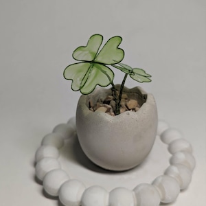 Op de afbeelding: Een kleine, doorschijnende lichtgroene klaverbladsculptuur van draad en hars, geplant in een grijze, eivormige betonnen pot gevuld met kleine steentjes, rustend in een cirkelvormige ring van witte kralen met een gladde, glanzende afwerking.