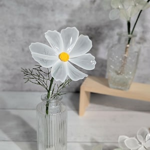 Könnte beinhalten: Eine einzelne künstliche weiße Kosmeenblüte mit leuchtend gelber Mitte, durchscheinenden Blütenblättern und einem dünnen grünen Stiel mit zartem Blattwerk, präsentiert in einer klaren, vertikal gerippten Glasflaschenvase.