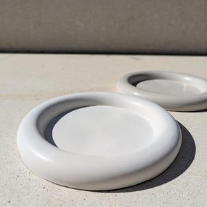 Pode incluir: Dois porta-copos redondos e minimalistas. Um é branco, o outro cinza claro. Ambos têm uma borda arredondada e elevada e um centro plano. Os porta-copos são feitos de um material liso e sólido.