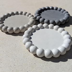 Puede incluir: Tres posavasos redondos y decorativos en tonos grises y blancos. Cada posavasos tiene un centro liso y plano y un borde con cuentas. Los posavasos están dispuestos sobre una superficie clara y texturizada.