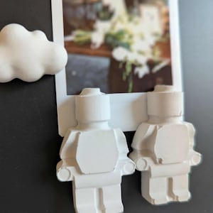 以下が含まれることがあります： レゴのフィギュアのような、白いセラミック製の人の置物が2つ、黒い表面に付けられています。その上に白い雲の置物があり、後ろには白い花のブーケの写真があります。