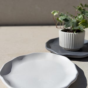 Può includere: Un piatto grigio chiaro, di forma organica, con un bordo ondulato. Un piatto più piccolo, grigio scuro, contiene un vaso bianco e scanalato con una piccola pianta verde. La scena è illuminata dalla luce solare.
