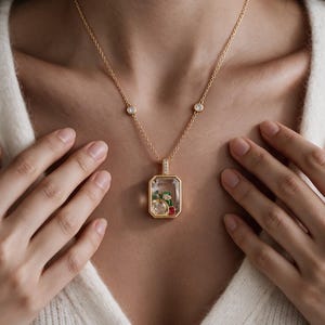 May include: A gold-tone necklace featuring a rectangular pendant with a clear glass casing. Inside the pendant, there are small, colourful floral and fruit-shaped charms in red, green, and white. The chain includes two small, round, clear stone accents.